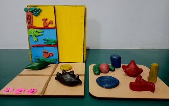 The StoryBox prototype: a yellow wooden box with tactile clay paintings of a monkey and crocodiles, an interactive mat with braille-numbered buttons, and a tray of clay figurines.